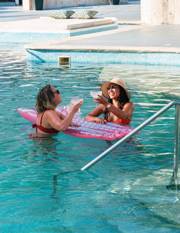 Two women chatting with drinks and a pink float in a turquoise resort pool.