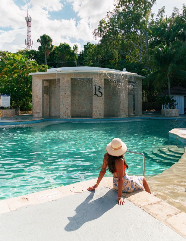 Woman wearing a sunhat sits at the pool edge facing a circular waterfall feature.