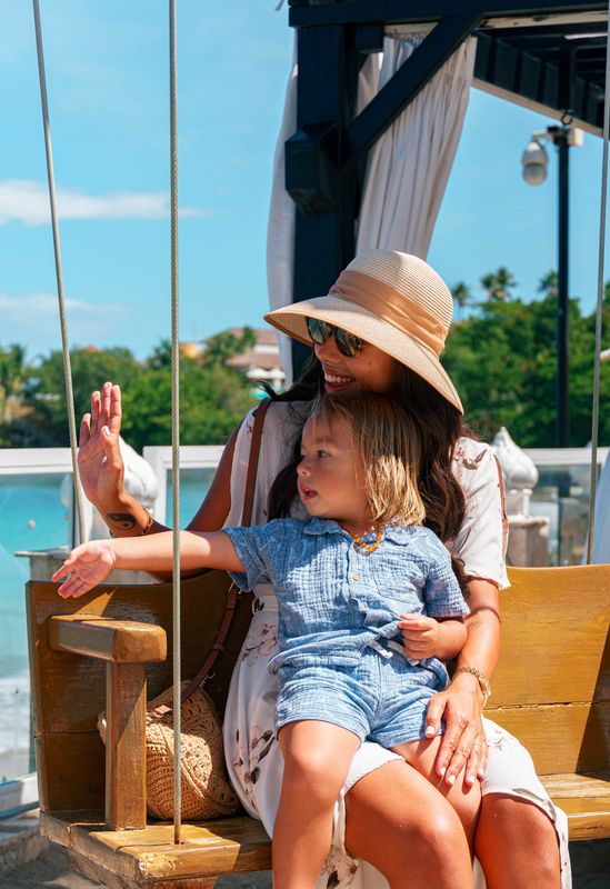 Woman in a sunhat and child in blue sit together on a wooden seaside swing.