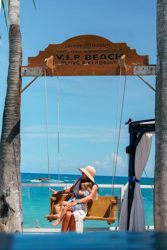 Woman and child on a beach swing beneath a VIP Beach sign, with the sea beyond.