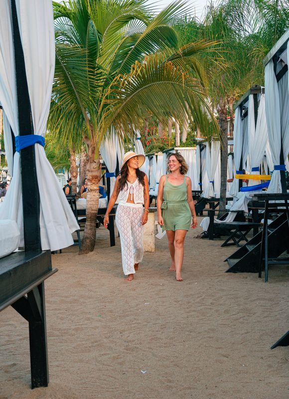 Two women walk along a sandy palm-lined path between curtained beach cabanas.