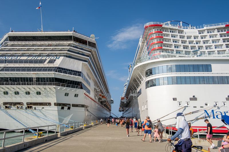 Passengers walk along a pier between two large docked cruise ships.
