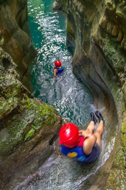 Overhead view of a red-helmeted visitor sliding through a narrow rock channel into turquoise water.
