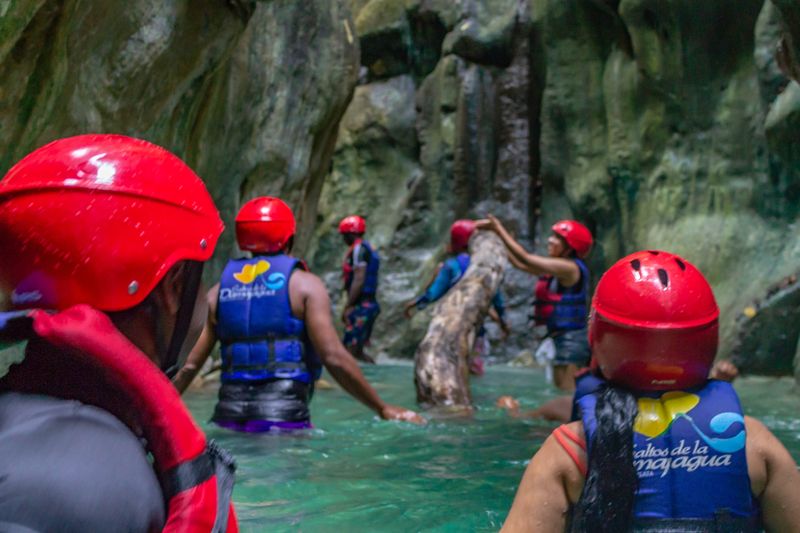Visitors wearing red helmets and blue life jackets wade through a rocky canyon pool.