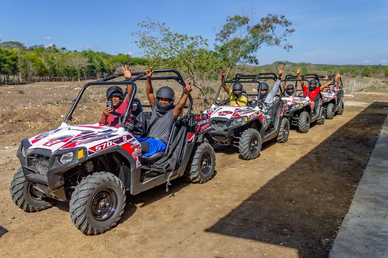 A line of red-and-white off-road buggies with riders on a sunny dirt track.