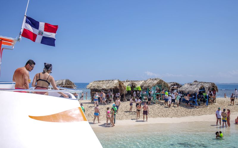 Boat with a Dominican flag approaches a sandy islet with thatched shelters and beachgoers.
