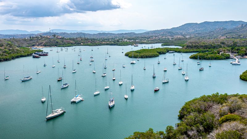 Aerial view of sailboats anchored in a sheltered bay beside green islets and distant hills.