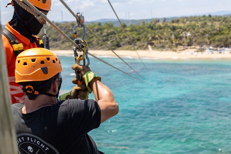 Helmeted visitor prepares for a zipline above turquoise coastal water with an instructor nearby.