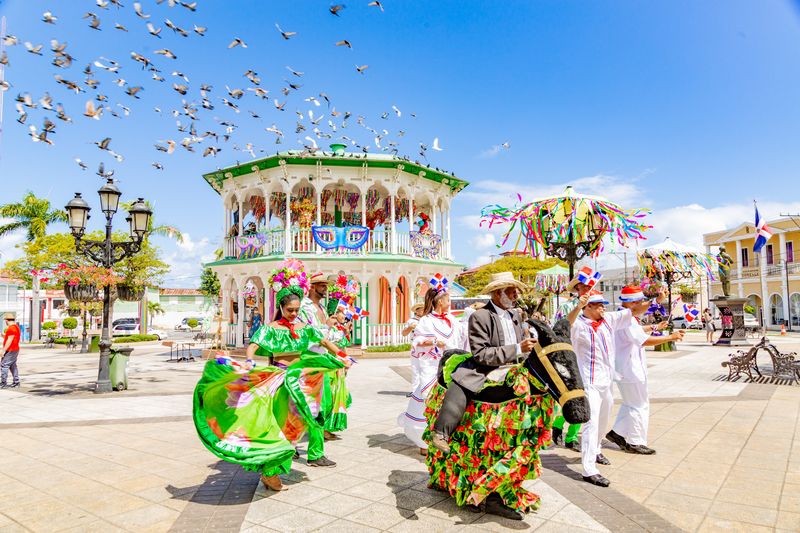 Dancers in green and white costumes perform in a sunny square as birds fly above the gazebo.