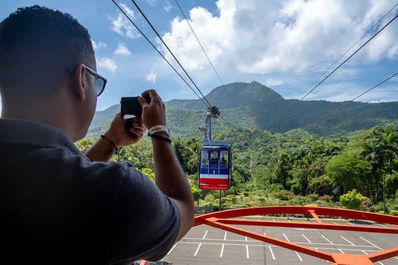 Visitor photographs a red cable car traveling above lush green mountains.