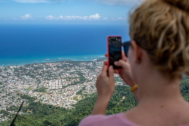 Visitor uses a phone to photograph a coastal city and blue ocean from a high viewpoint.