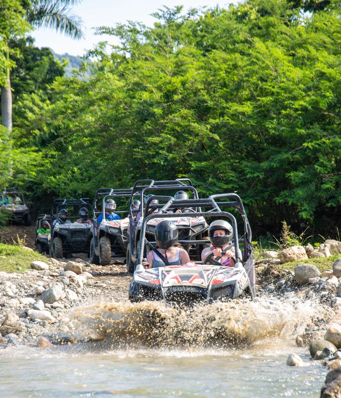 Off-road buggies splash through a rocky stream surrounded by tropical forest.